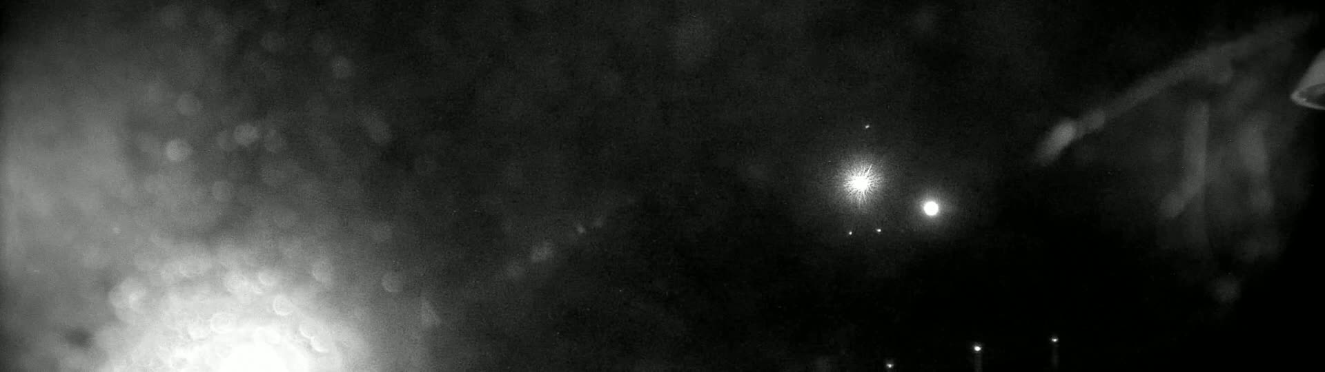 A blurry, nighttime image shows several bright lights, possibly distant lights or stars, amidst a dark and hazy sky.
