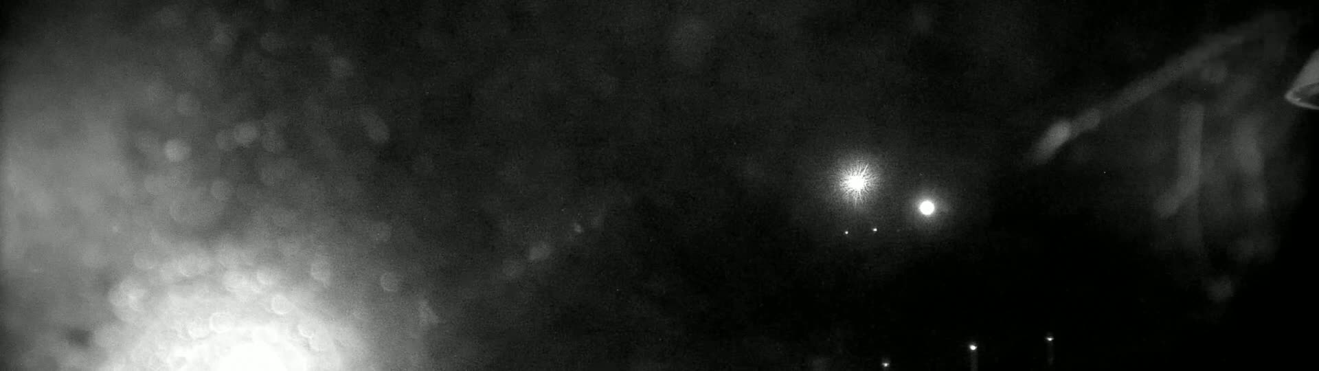 A dark, blurry night scene shows bright lights in the distance, possibly streetlights, through a hazy or rainy atmosphere.