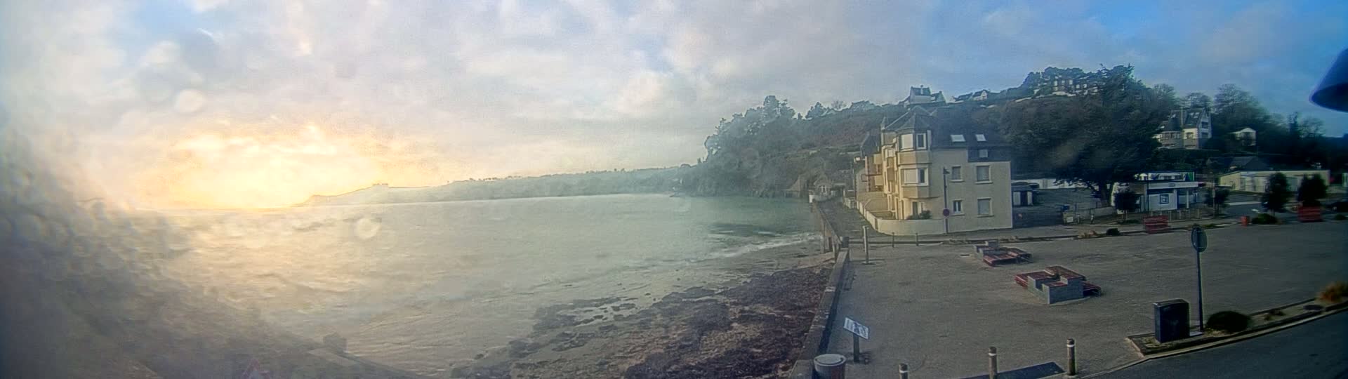 A seaside town at sunrise shows a calm body of water, a rocky shoreline, and buildings on a hillside under a partly cloudy sky.