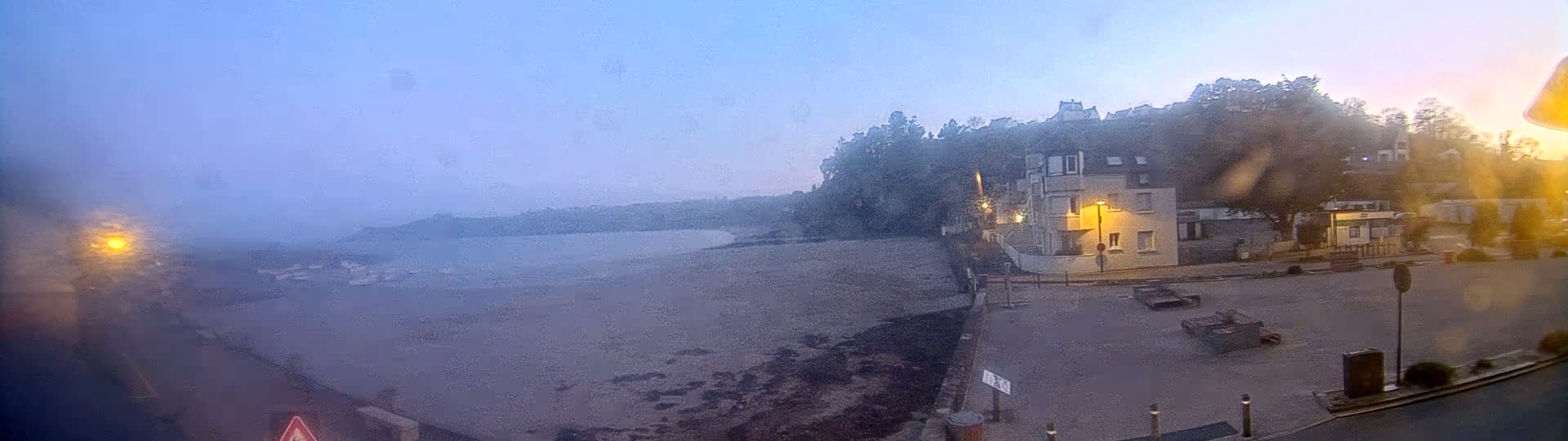 A foggy, twilight view of a calm body of water, a sandy beach, and buildings on a hillside.