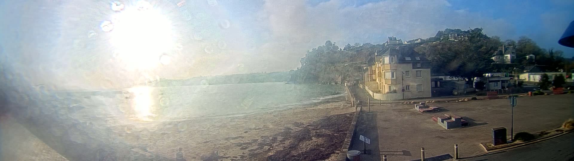 A sunny, partly cloudy day shows a sandy beach, calm water, and a multi-story building near a hillside with trees and other houses.