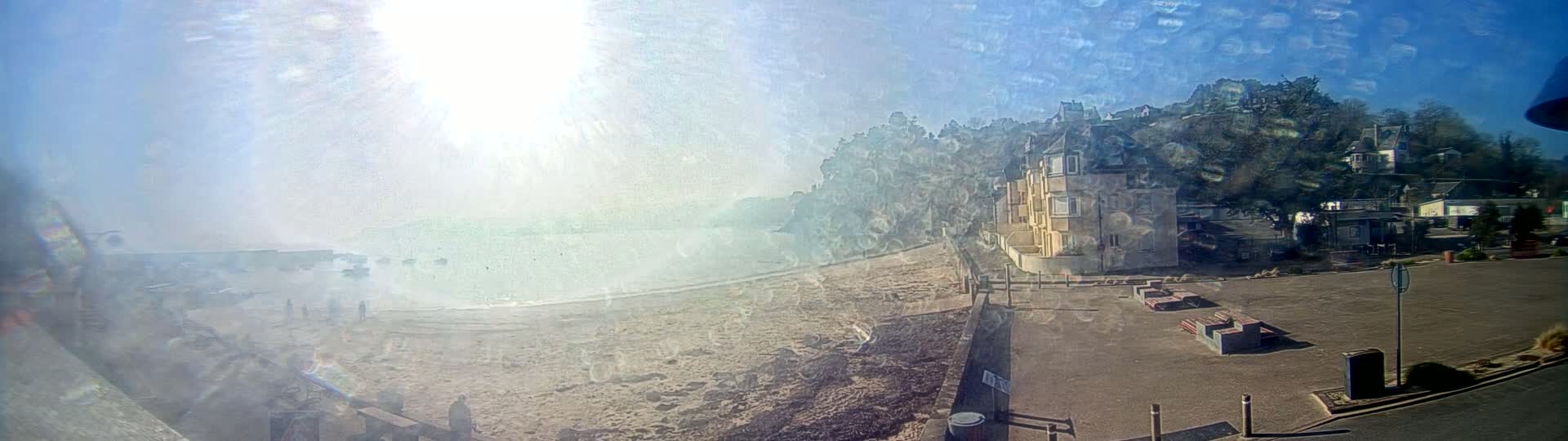 A hazy, sunny day reveals a sandy beach, calm water, and a building overlooking the scene from a hillside, with a parking lot in the foreground.