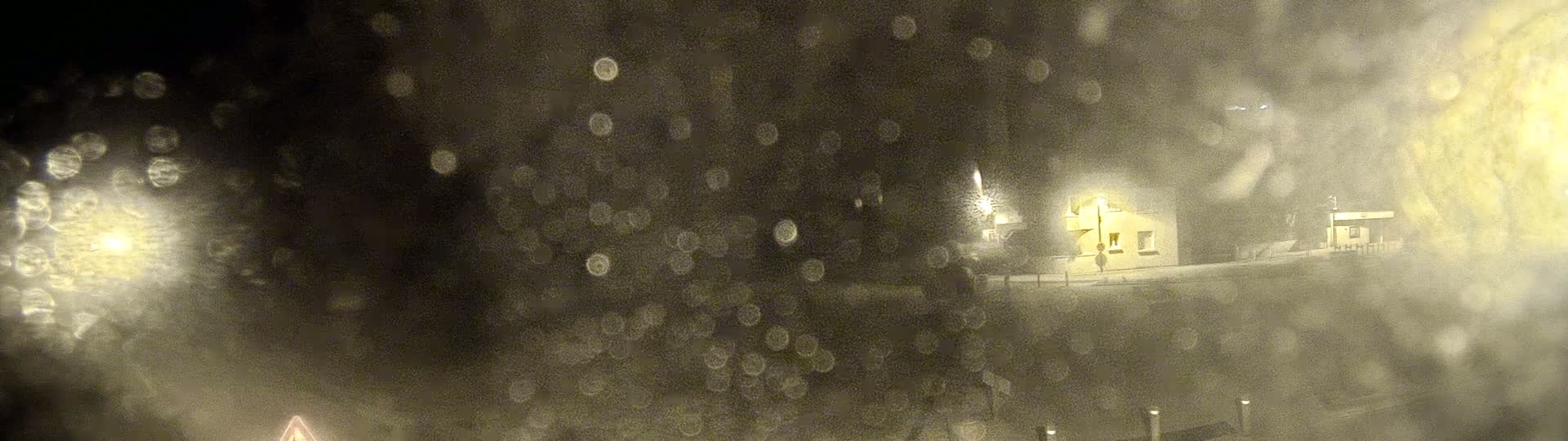 A blurry nighttime view through rain-covered glass shows a small building faintly illuminated by a streetlight.