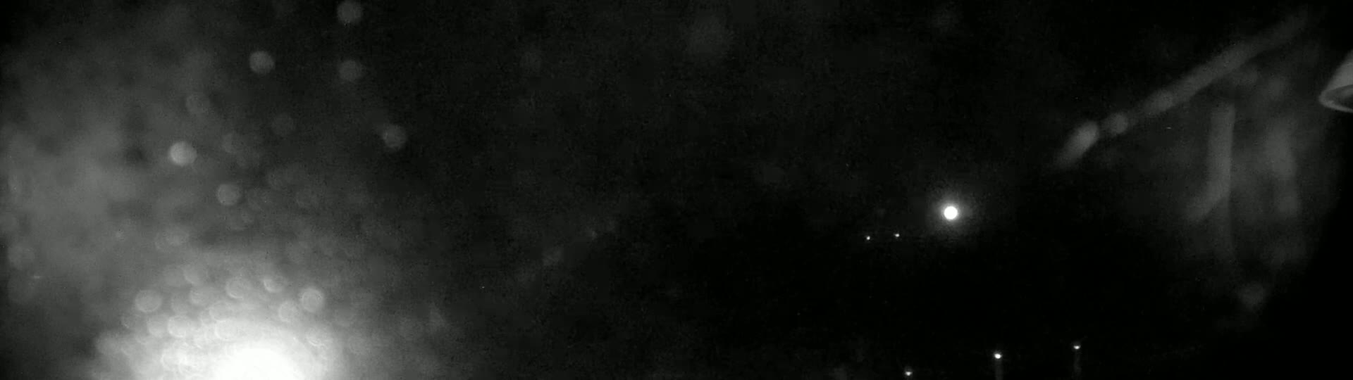 A nighttime, rainy scene shows blurry lights in the distance and rain drops on a dark surface.