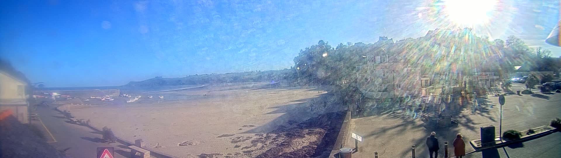 A sunny day reveals a sandy beach with small boats, a seaside town with buildings and trees, and a few people walking near the water.