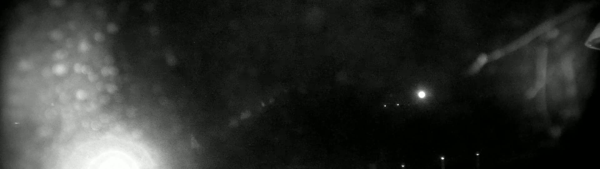 A blurry, nighttime image shows bright lights and a bright, possibly moonlit sky, with some rain or snow visible.