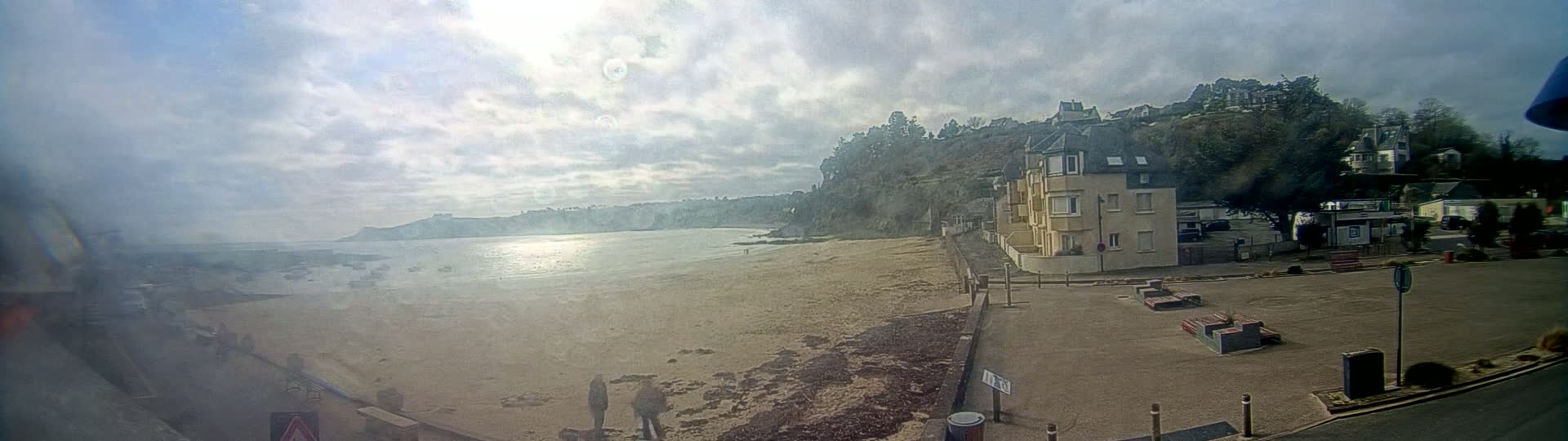 A sandy beach with calm water and a few buildings on a hillside under a partly sunny sky.