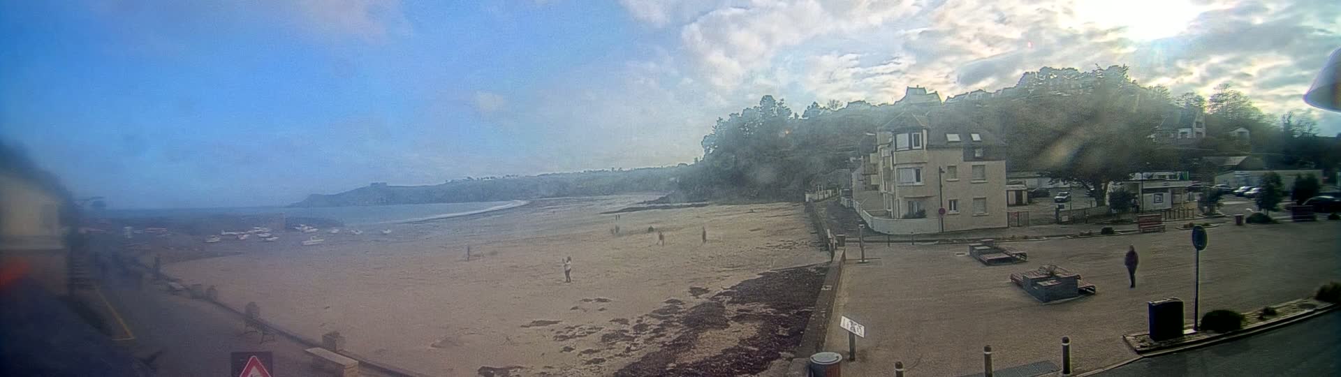 A sandy beach stretches out to a calm ocean under a partly cloudy sky, with a few people scattered on the beach and a building and parking lot visible nearby.