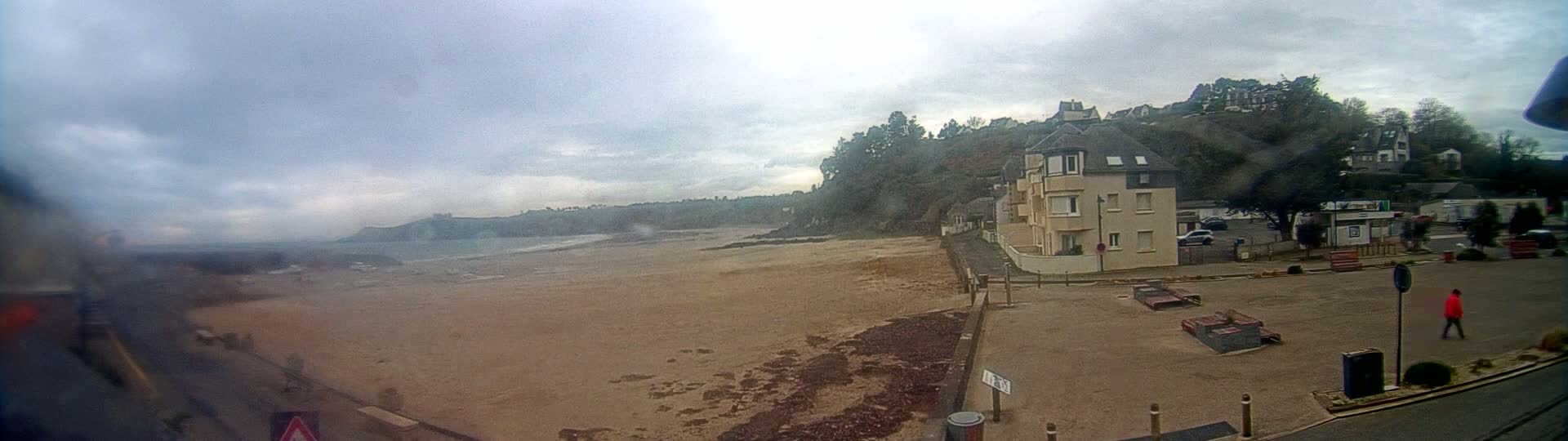 A sandy beach stretches out to a calm ocean under a cloudy sky, with buildings and trees on a hill overlooking the shore.