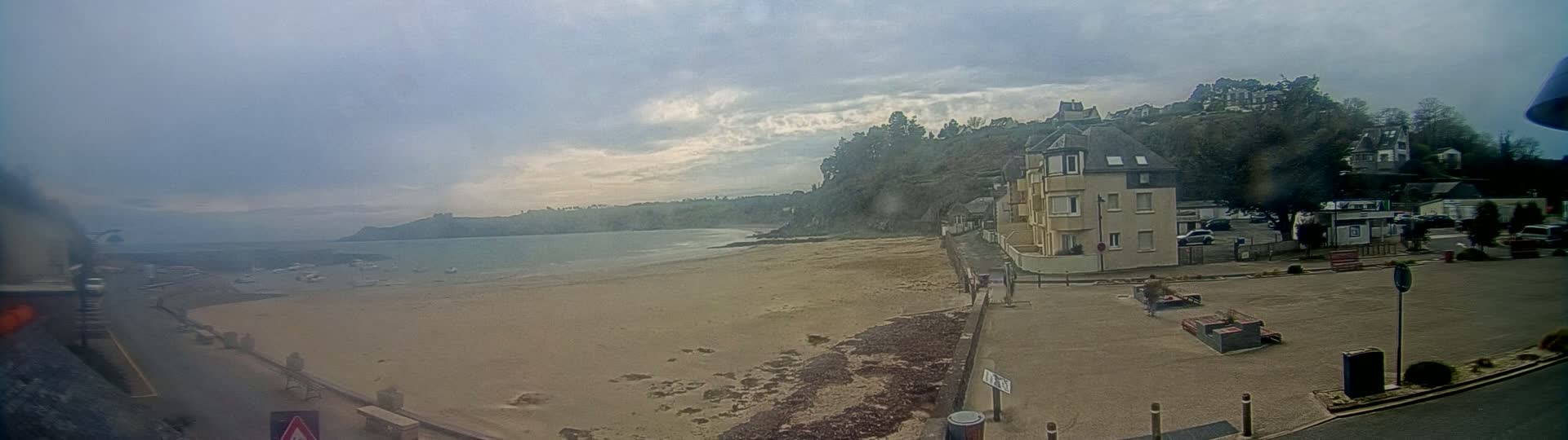 A sandy beach stretches out to a calm body of water under a cloudy sky, with buildings and trees rising on a hillside in the background.