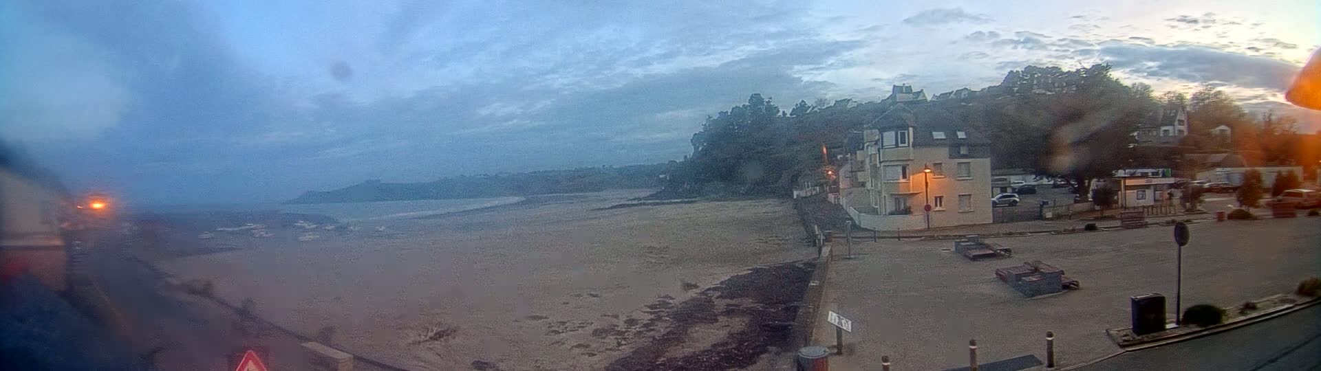 A sandy beach stretches out to a calm ocean under a cloudy, dusky sky, with a building and parked cars visible in the foreground.