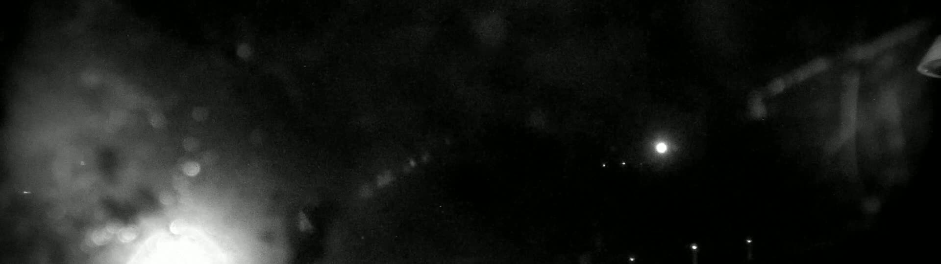 A blurry, nighttime image shows bright lights and what appears to be rain or snow falling in front of a dark background.