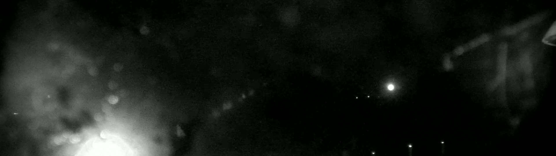 A blurry nighttime image shows bright lights and dark shapes, possibly buildings, in the distance, with a hazy or rainy atmosphere.