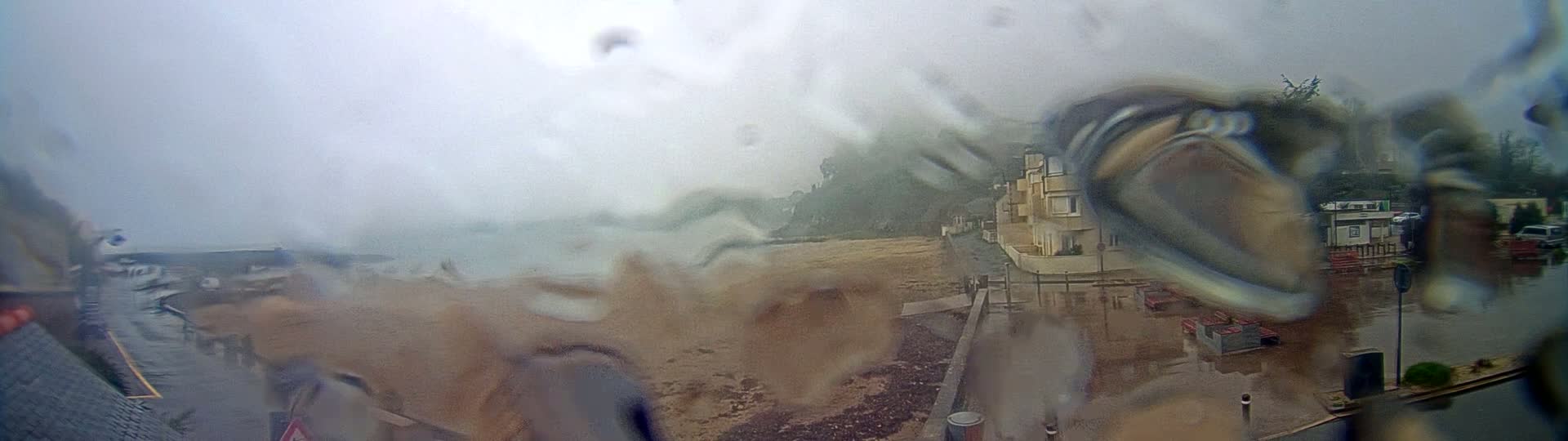 A rain-streaked view shows a coastal scene with buildings and a beach partially submerged in floodwater under an overcast sky.