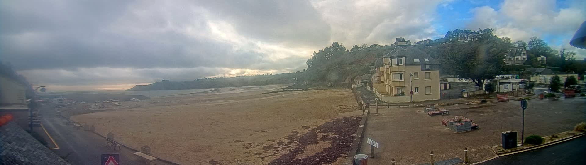 A sandy beach stretches out to a calm sea under a cloudy sky, with buildings and trees on a hillside in the background.