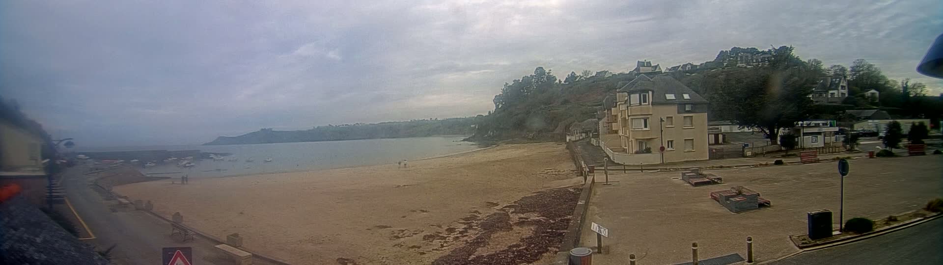 A sandy beach next to a calm bay is overlooked by a building and houses on a hillside under an overcast sky.