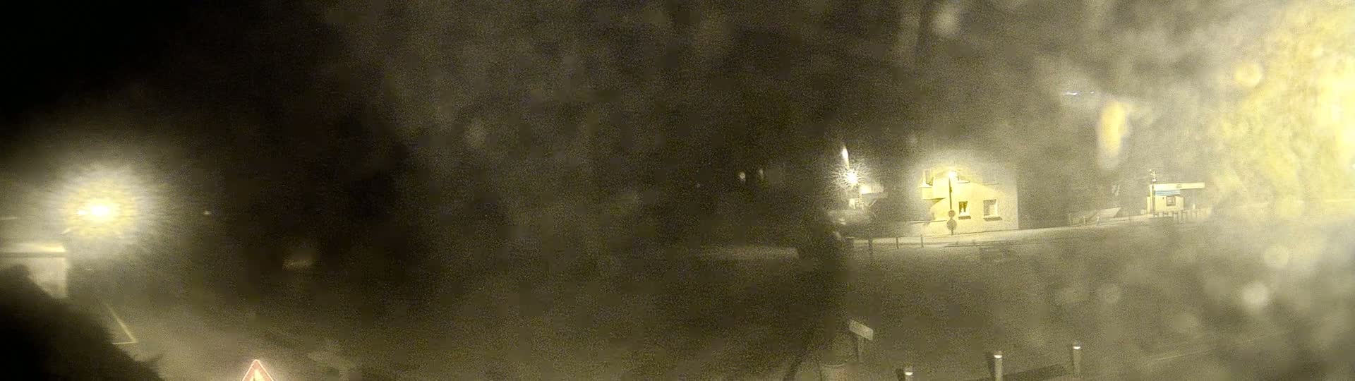 A dimly lit, snow-covered scene at night shows a small building and streetlights through a blurry, snow-covered window.