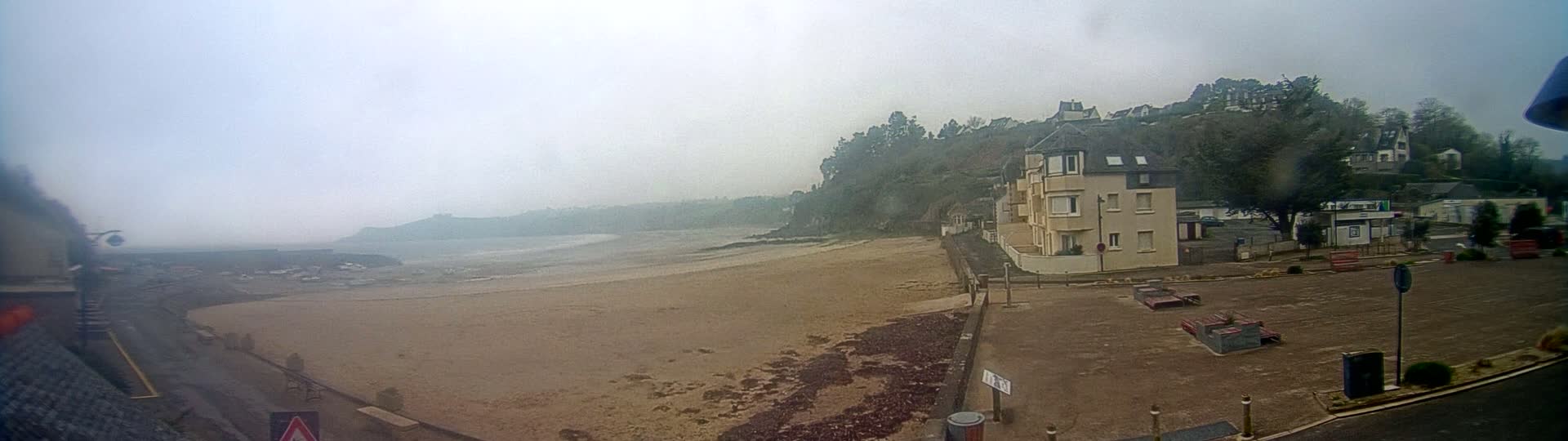 A sandy beach stretches out to a misty ocean under an overcast sky, with buildings and trees rising on a hill behind it.