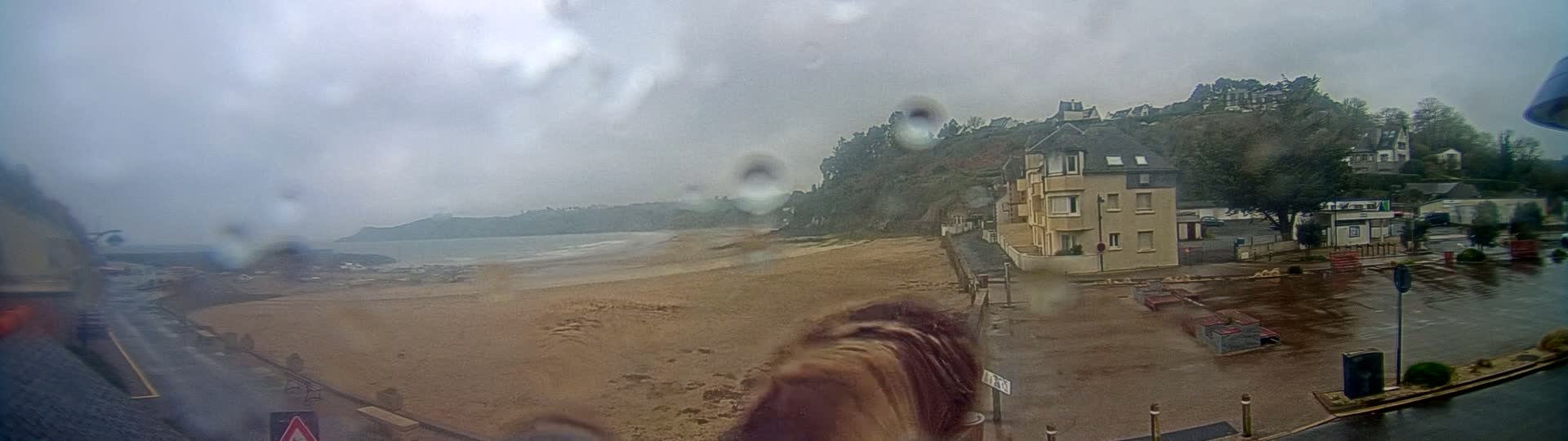 A rain-swept view of a sandy beach, calm ocean, and a seaside town with buildings on a hillside, seen through a rain-covered window.