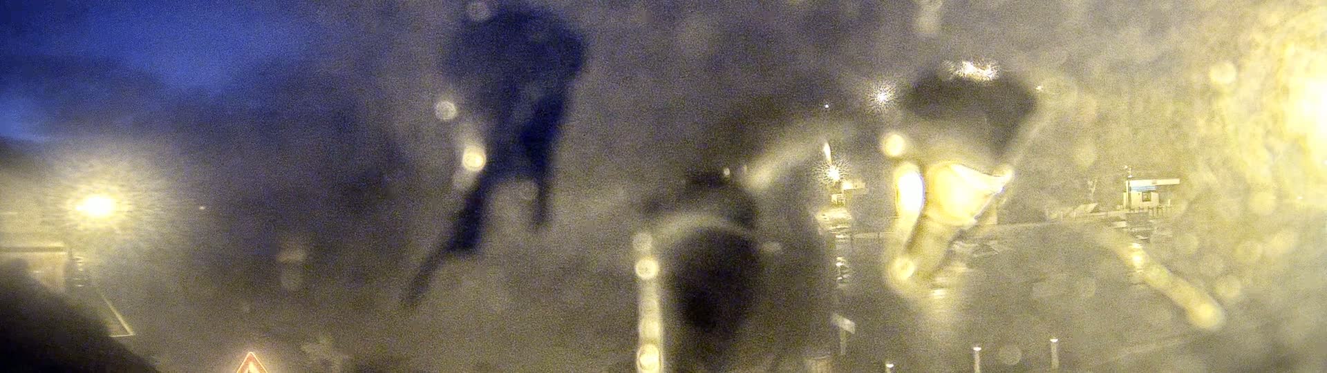 Blurry figures are seen through a rain-streaked window at night, illuminated by streetlights.