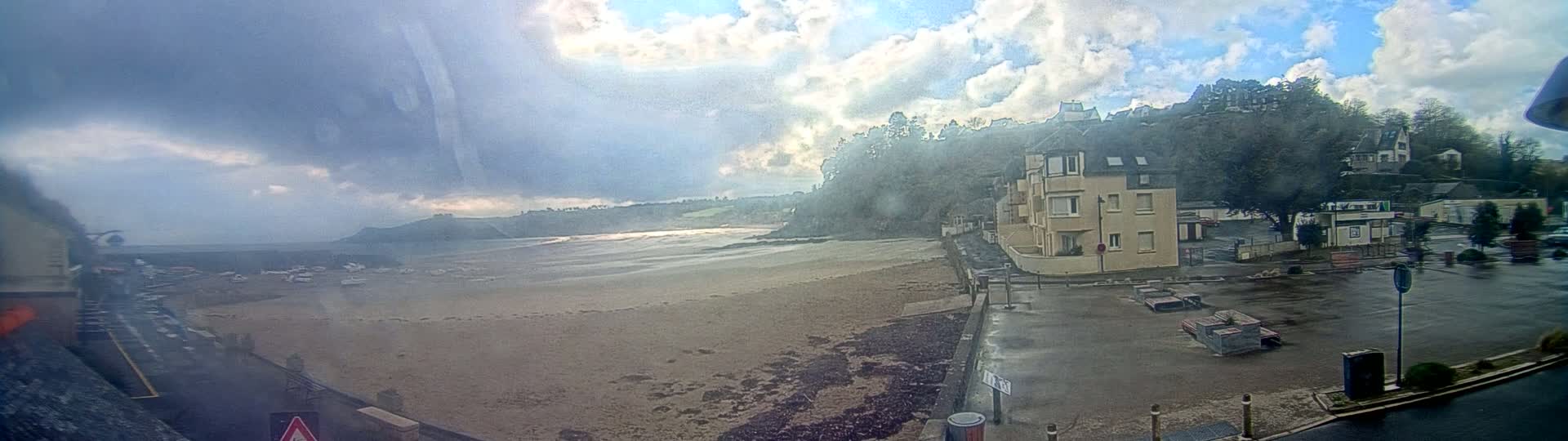 A mostly cloudy, rainy day reveals a sandy beach, a calm body of water, and buildings situated on a hillside overlooking the coast.