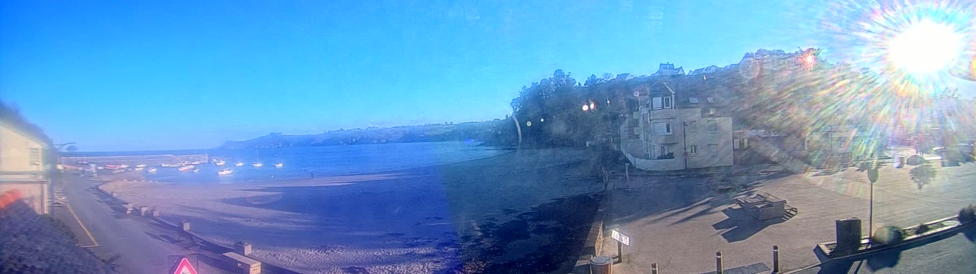 A sandy beach with several small boats in the water is visible below a hillside with buildings, all under a bright, sunny sky.