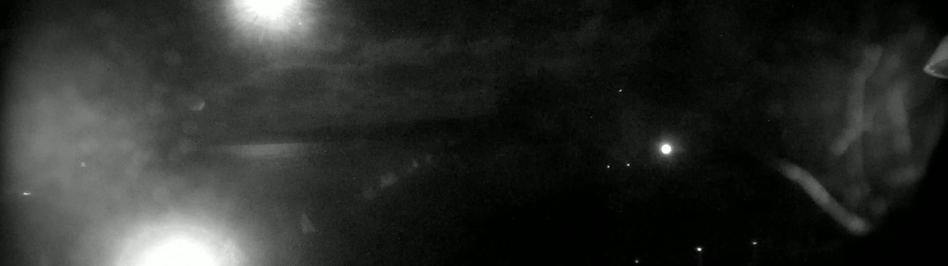 A nighttime, hazy or foggy scene shows indistinct shapes and bright lights, possibly from streetlights or headlights.