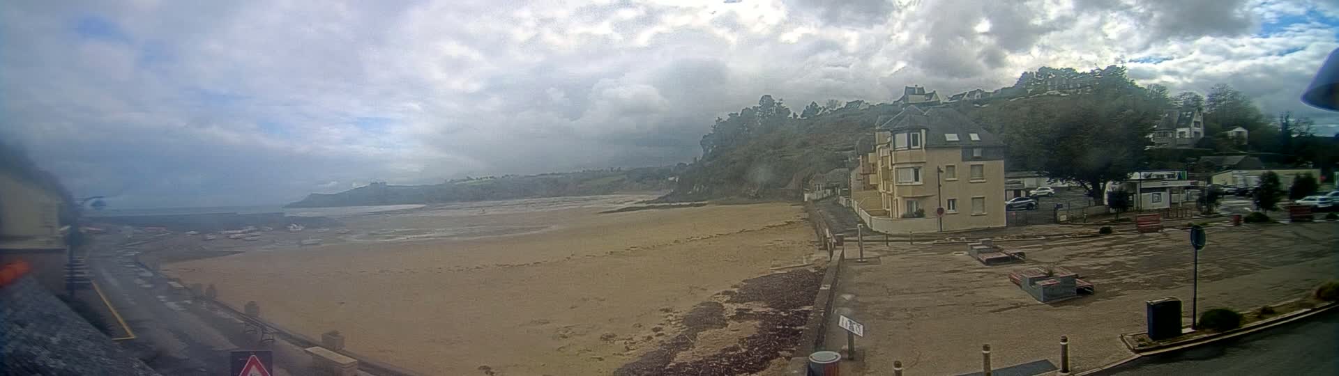 A sandy beach stretches out to a calm sea under a cloudy sky, with buildings and trees rising on a hillside behind it.
