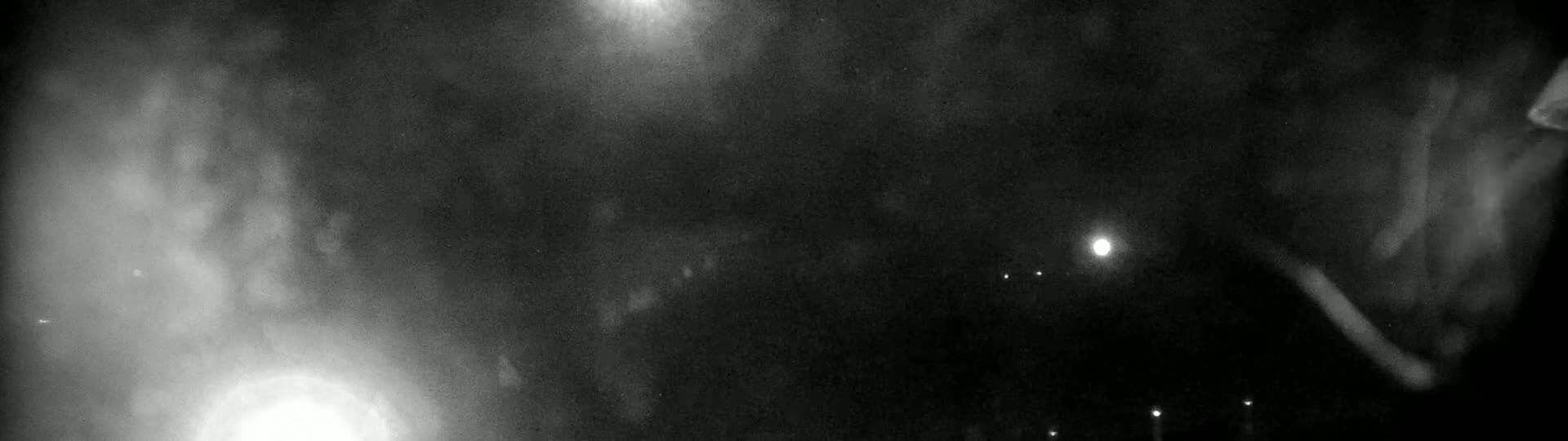 A blurry, grainy night-time image shows bright lights and faint shapes in the darkness, possibly due to fog or poor visibility.