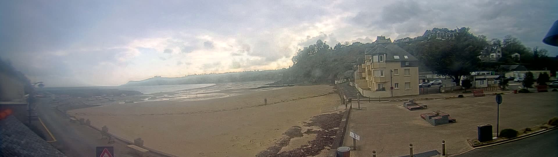 A sandy beach stretches out to a calm ocean under a cloudy sky, with buildings and trees lining a hillside in the background.