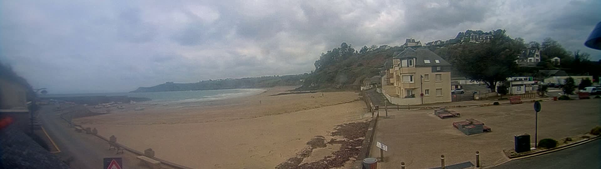 A sandy beach with gentle waves stretches out under an overcast sky, backed by a hillside with buildings and trees.