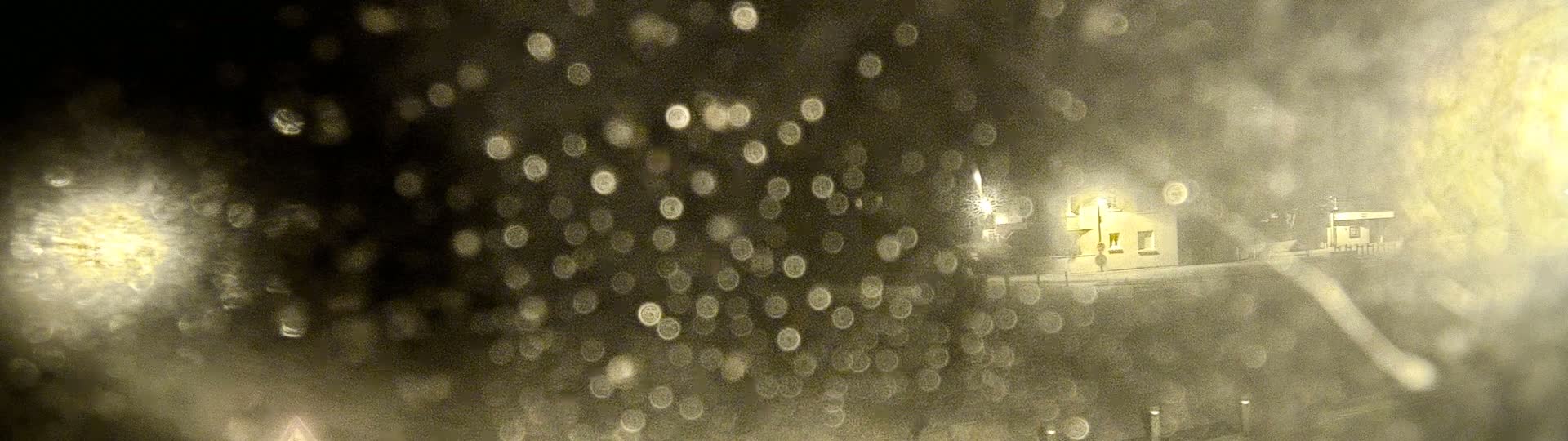 A blurry, nighttime view of a building through a rain-covered window, showing out-of-focus lights and water droplets.
