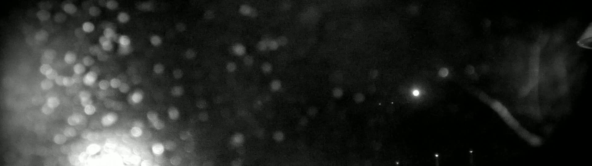 A blurry, black-and-white nighttime image shows out-of-focus lights and what appears to be rain or snow falling.