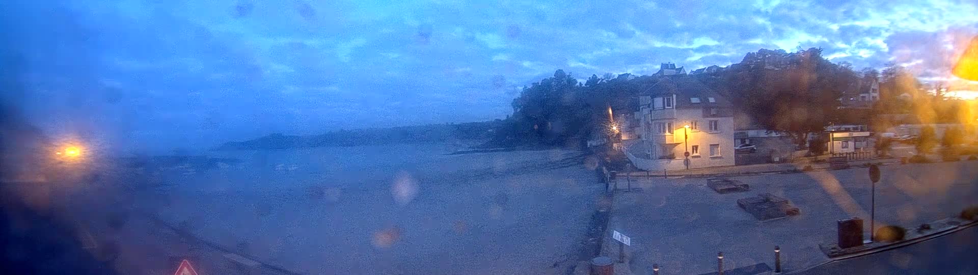 A coastal town scene at twilight under a cloudy sky shows a building near a mostly empty parking lot overlooking a calm body of water.