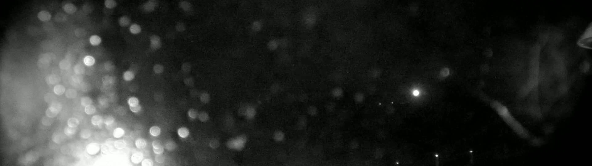 A nighttime, blurry image shows out-of-focus lights, possibly from rain or snow, with a few distant, brighter lights visible in the background.