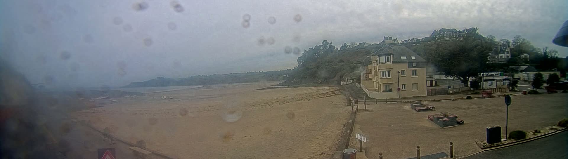 A rain-streaked view shows a sandy beach, a few boats in the water, and a tan apartment building overlooking the scene, all under an overcast sky.