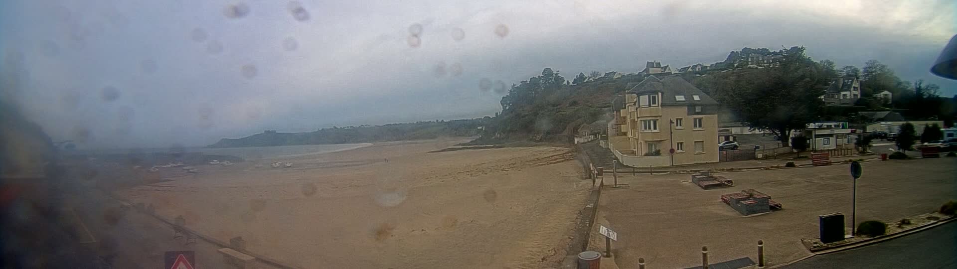 A sandy beach stretches to a calm sea under an overcast sky, with a building and parking lot visible nearby.