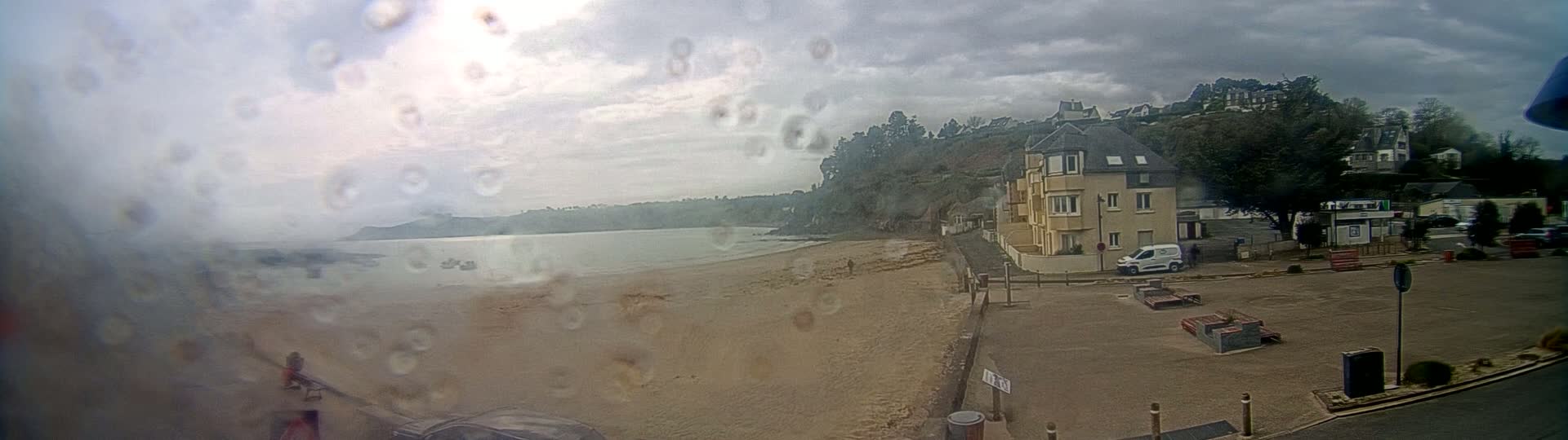 A rain-covered view shows a sandy beach, calm water, buildings on a hillside, and a parking lot with some park benches.