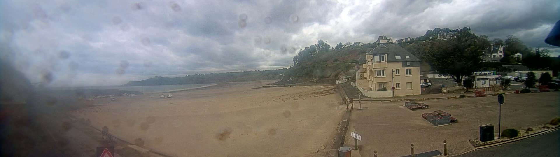 A sandy beach stretches out under a cloudy sky, with a multi-story building and a parking area visible closer to the viewer.