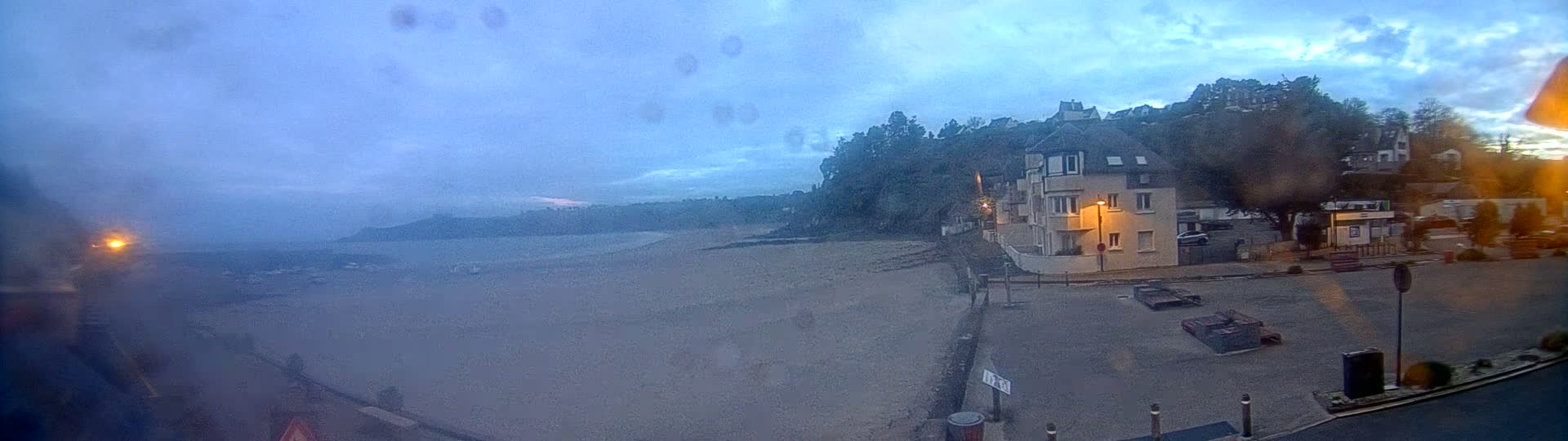 A mostly empty sandy beach stretches out under a dusky, cloudy sky, with a building and some trees situated on a cliff overlooking it.