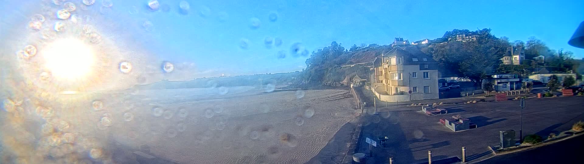 A sandy beach stretches out to a calm ocean under a bright, sunny sky, viewed through a rain-covered window, with a building and parking lot visible on the right.
