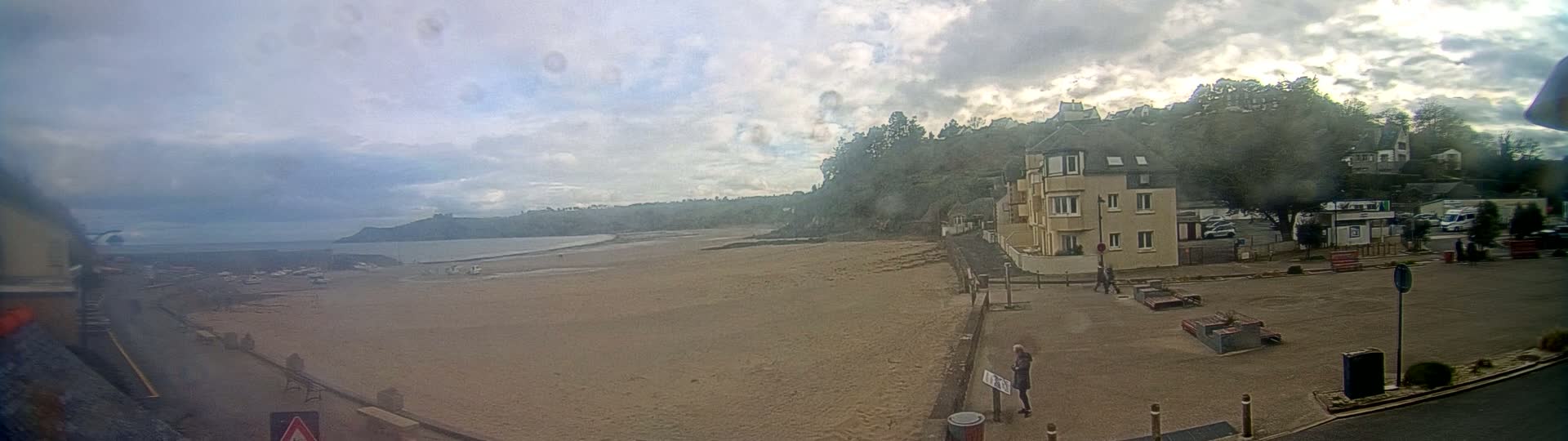 A wide shot of a sandy beach with a few people visible, an apartment building, and a parking lot on an overcast day.