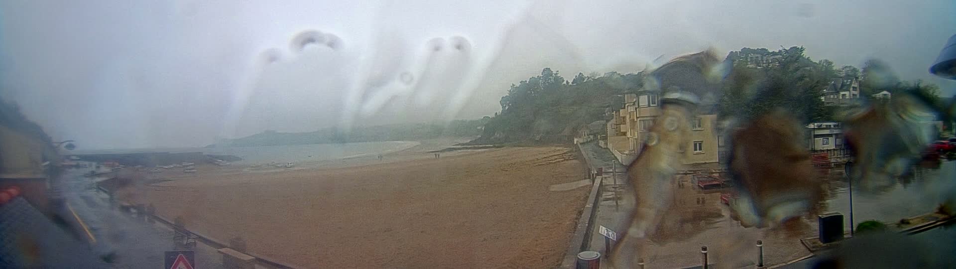 A rain-blurred view shows a sandy beach, a calm body of water, buildings along a shoreline, and a hill covered in trees in the background.