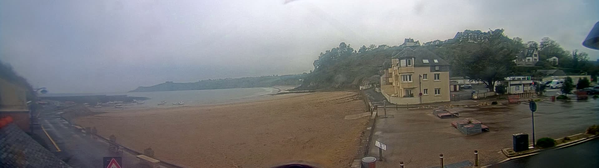 A sandy beach with a few boats is visible on a foggy day, next to a parking lot with a multi-story building and other buildings on a hillside behind it.