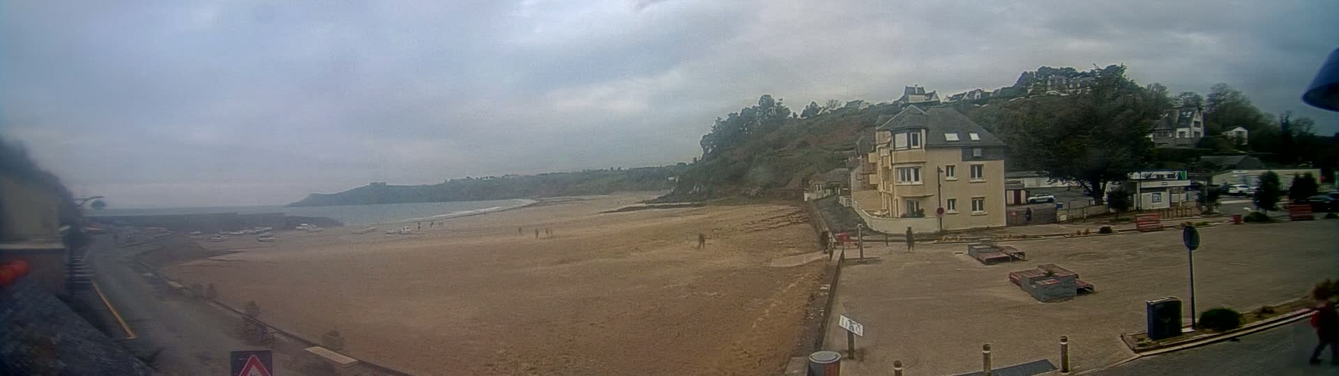 A sandy beach stretches out to a calm ocean under an overcast sky, with a few people scattered across the sand and a multi-story building overlooking the scene from a nearby hill.