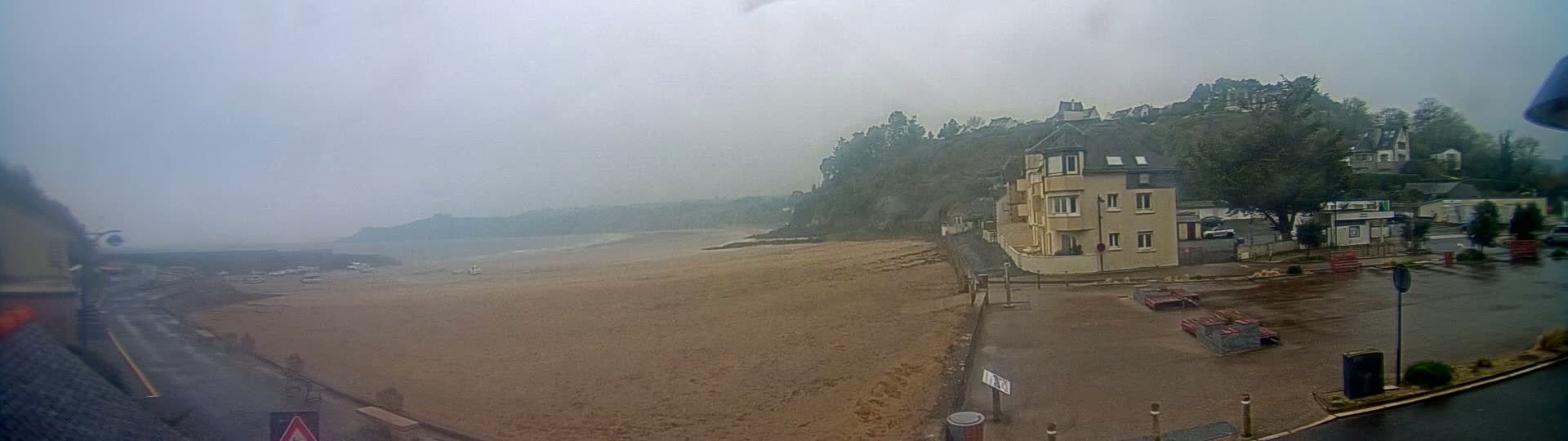 A sandy beach stretches out to a hazy ocean under an overcast sky, with a building and parking lot visible near a tree-covered hillside.
