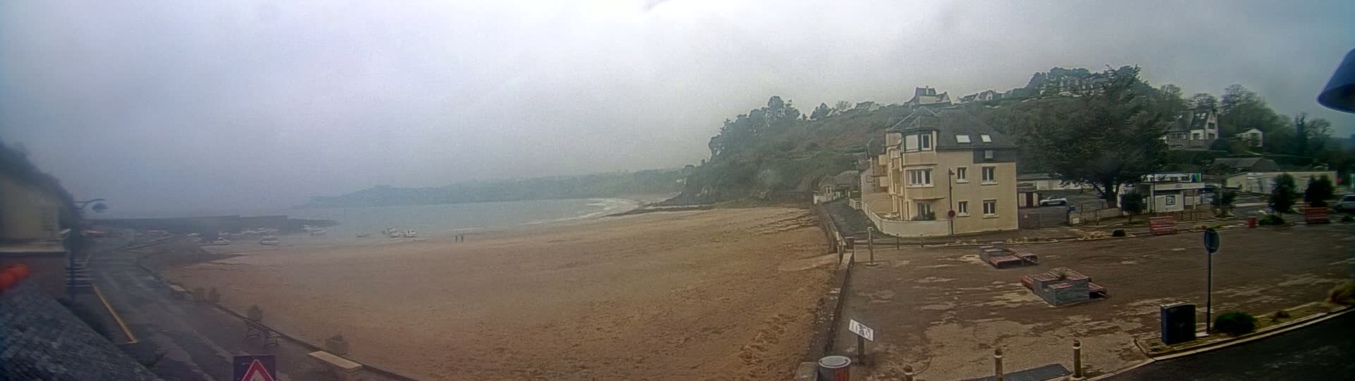 A sandy beach stretches out to a calm, foggy sea under an overcast sky, with buildings and greenery on a hillside overlooking the scene.