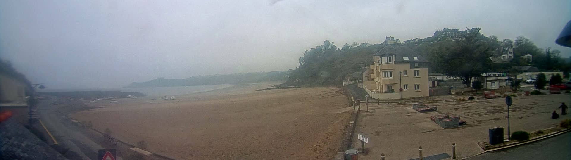 A foggy coastal scene shows a sandy beach, a calm body of water, and a light beige building near a hill covered in trees.