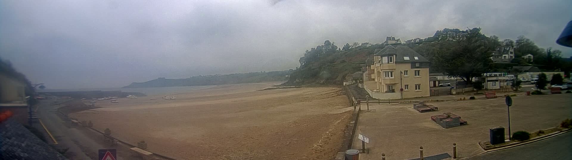 A sandy beach stretches to a calm, gray sea under an overcast sky, with a multi-story building and parking lot situated on a grassy hill overlooking the shore.