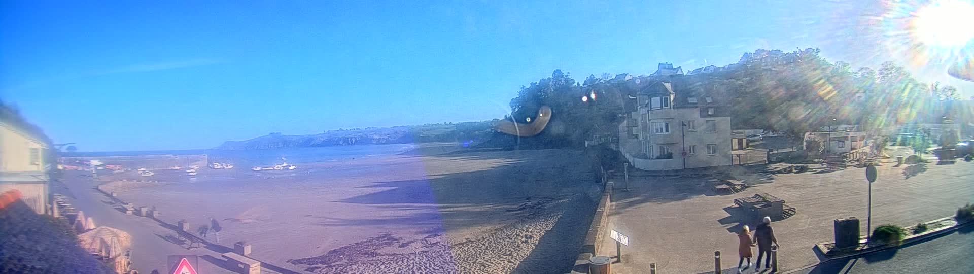 A sandy beach with boats in the water is overlooked by buildings and green hills under a sunny sky.